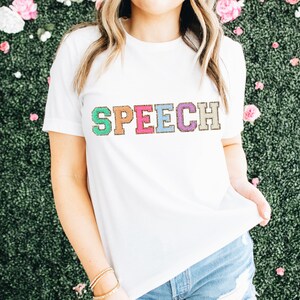 May include: White t-shirt with the word "SPEECH" in colorful, glittery letters.