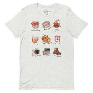 May include: A light gray t-shirt with a design of nine fall-themed illustrations. The illustrations include apple picking, baking pies, roasting marshmallows, pumpkin spice, pumpkin carving, fall candles, family photos, movie marathon, and nature hikes.