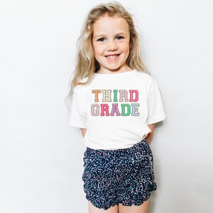 May include: A white t-shirt with the words "THIRD GRADE" in colorful letters. The letters are outlined in a light brown color. The shirt is being worn by a young girl with long blonde hair.