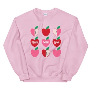 Puede incluir: Sudadera rosa con un gr&aacute;fico de manzanas rojas y rosas y el texto "Teach with Love".