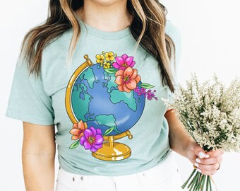 Puede incluir: Una camiseta azul turquesa con un gráfico de un globo terráqueo rodeado de flores de colores. El globo es dorado y azul con continentes verdes.