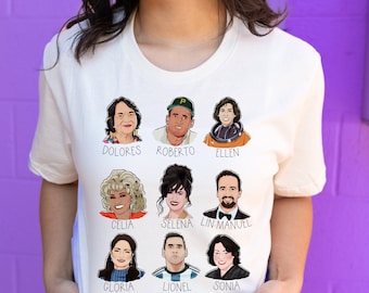 Op de afbeelding: Wit T-shirt met een raster van negen cartoonportretten van mensen, elk met een naam eronder. De namen zijn: Dolores, Roberto, Ellen, Celia, Selena, Lin-Manuel, Gloria, Lionel, Sonia.