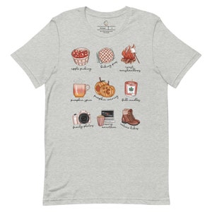 May include: A gray t-shirt with a white design of fall activities. The design includes illustrations of apple picking, baking pies, roasting marshmallows, pumpkin spice, pumpkin carving, fall candles, family photos, movie marathon, and nature hikes.