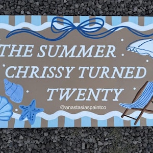May include: A rectangular wooden sign with the text "THE SUMMER CHRISSY TURNED TWENTY" in white. The sign features a beach theme with blue seashells, a starfish, and a beach chair. The top has a blue bow.