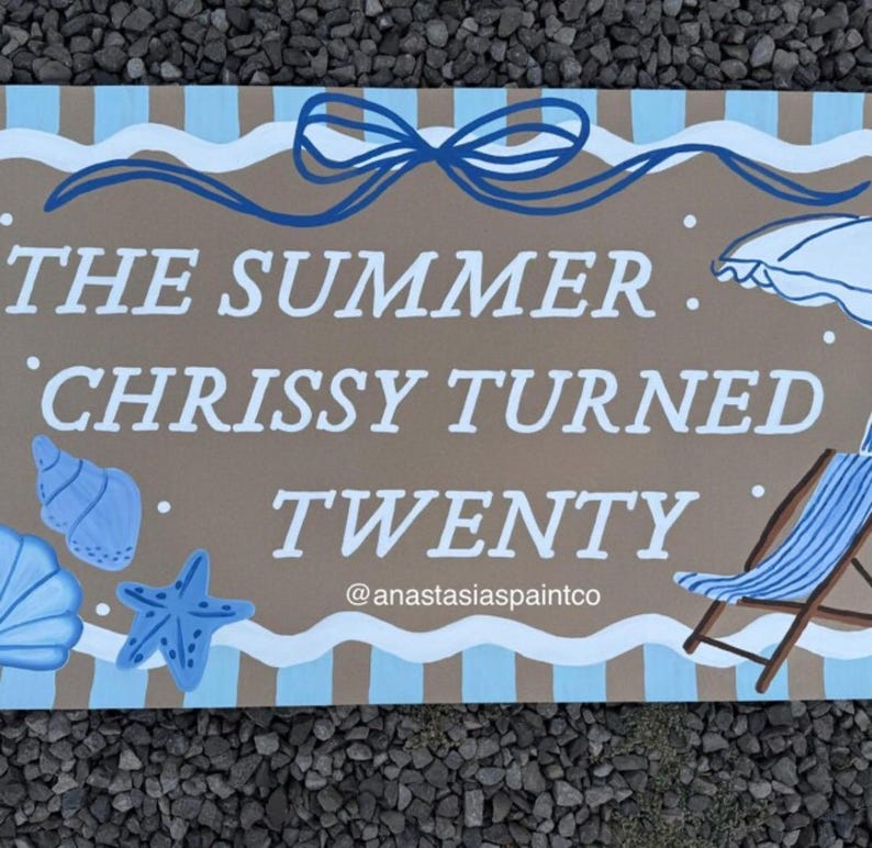 May include: A rectangular wooden sign with the text "THE SUMMER CHRISSY TURNED TWENTY" in white. The sign features a beach theme with blue seashells, a starfish, and a beach chair. The top has a blue bow.