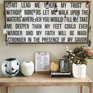 May include: A wooden sign with the text "Spirit Lead Me Here My Trust Without Borders Let Me Walk Upon The Waters Wherever You Would Call Me Take Me Deeper Than My Feet Could Ever Wander And My Faith Will Be Made Stronger In The Presence Of My Savior". The sign is decorated with a string of lights and small photos.
