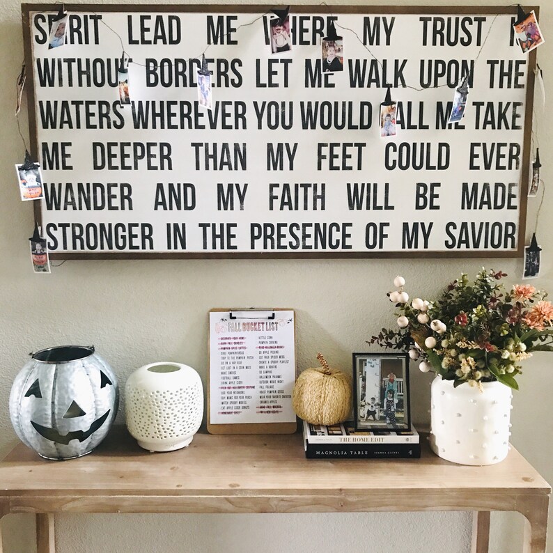 May include: A wooden sign with the text "Spirit Lead Me Here My Trust Without Borders Let Me Walk Upon The Waters Wherever You Would Call Me Take Me Deeper Than My Feet Could Ever Wander And My Faith Will Be Made Stronger In The Presence Of My Savior". The sign is decorated with a string of lights and small photos.