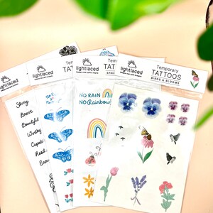 May include: Four sheets of temporary tattoos with various designs. The first sheet features the words "Strong, Brave, Beautiful, Worthy, Capable, Resilient, Enough". The second sheet features a rainbow with the words "No Rain No Rainbow". The third sheet features a butterfly and the words "No Rain No Rainbow". The fourth sheet features a variety of flowers, birds, and butterflies with the words "Temporary Tattoos Birds & Blooms".
