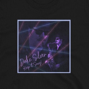Puede incluir: Camiseta negra con un gráfico morado y azul de un hombre tocando el saxofón. El gráfico tiene el texto "Duke Silver Keep it Savvy!" en morado.