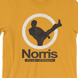 May include: A yellow t-shirt with a white circle and a black silhouette of a person kicking. The text "Norris Anti-Villain" is printed below the circle.