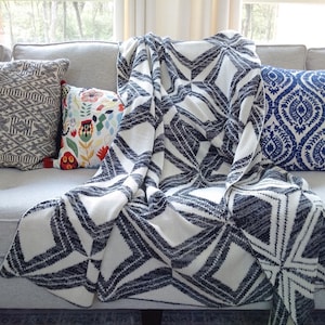 May include: A white and navy blue throw blanket with a geometric pattern is draped over a gray couch. The blanket is folded over the back of the couch and is partially covering the cushions. There are three pillows on the couch, one with a black and white geometric pattern, one with a colorful floral pattern, and one with a blue and white paisley pattern.