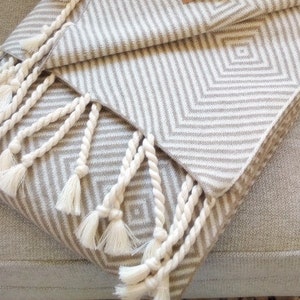 May include: A folded beige and white patterned blanket with diamond and chevron designs. The blanket has white tassels along the edge. The blanket is draped over a light gray upholstered chair.