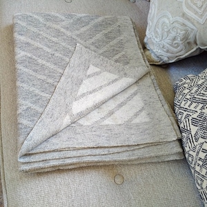 May include: A gray and white woven throw blanket folded on a light gray textured surface. The blanket has a subtle geometric pattern.