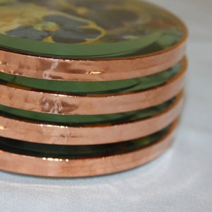 May include: A stack of four round glass coasters with copper trim. Each coaster features a pressed flower design.