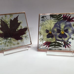 May include: Two square glass frames with copper trim, each containing pressed flowers and leaves. The frame on the left features a large, dark red maple leaf surrounded by small white flowers. The frame on the right features two purple and white pansies with other small flowers and leaves.