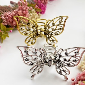 May include: Two butterfly-shaped hair clips, one in a gold-tone metal finish and one in a silver-tone metal finish. Each clip features an intricate, open-work filigree design with small floral-like details along the wings and a spring mechanism.