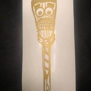 May include: Gold-coloured sticker featuring a stylised tiki design. The tiki has large, round eyes, a rectangular mouth, and a patterned body. The handle is striped. The sticker is on a white background.