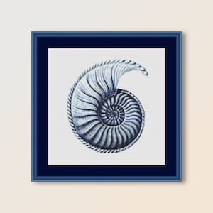 May include: A framed cross-stitch art piece featuring a detailed nautilus shell design in shades of blue and white. The shell is framed in a dark blue square frame, creating a nautical theme.