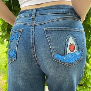 May include: Blue denim jeans with a shark painted on the back pocket. The shark is blue and white with a red mouth and white teeth. The words "Sharks are the friends" are written in blue on the left back pocket.