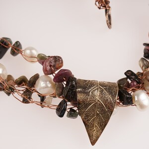 May include: A multi-strand necklace made with copper wire, small pink, green, and black stones, and white pearls. A gold leaf pendant hangs from the center of the necklace.