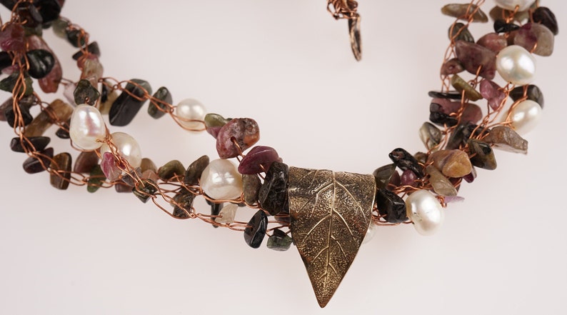 May include: A multi-strand necklace made with copper wire, small pink, green, and black stones, and white pearls. A gold leaf pendant hangs from the center of the necklace.