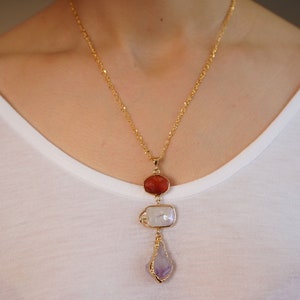 May include: A gold chain necklace with a pendant featuring three stones: a red carnelian, a white pearl, and a purple amethyst.