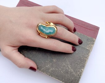 May include: A gold ring with a large, green, irregularly shaped stone set in the center. The ring has a textured, organic design.