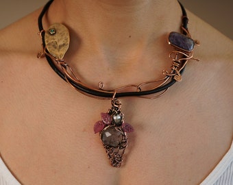 May include: A copper wire necklace with a black leather cord. The necklace features a pendant with a large gray gemstone, surrounded by smaller pink stones and a pearl. The necklace also has two other stones, one purple and one brown.