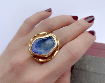 May include: A gold ring featuring a blue stone set in a hammered gold bezel. The stone is freeform in shape and has a rough, natural texture.