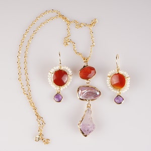 May include: A gold necklace and earrings set with red and purple gemstones. The necklace features a large, irregular-shaped purple stone, a smaller red stone, and a circle of white pearls. The earrings have a similar design with a red stone surrounded by white pearls and a smaller purple stone dangling below.