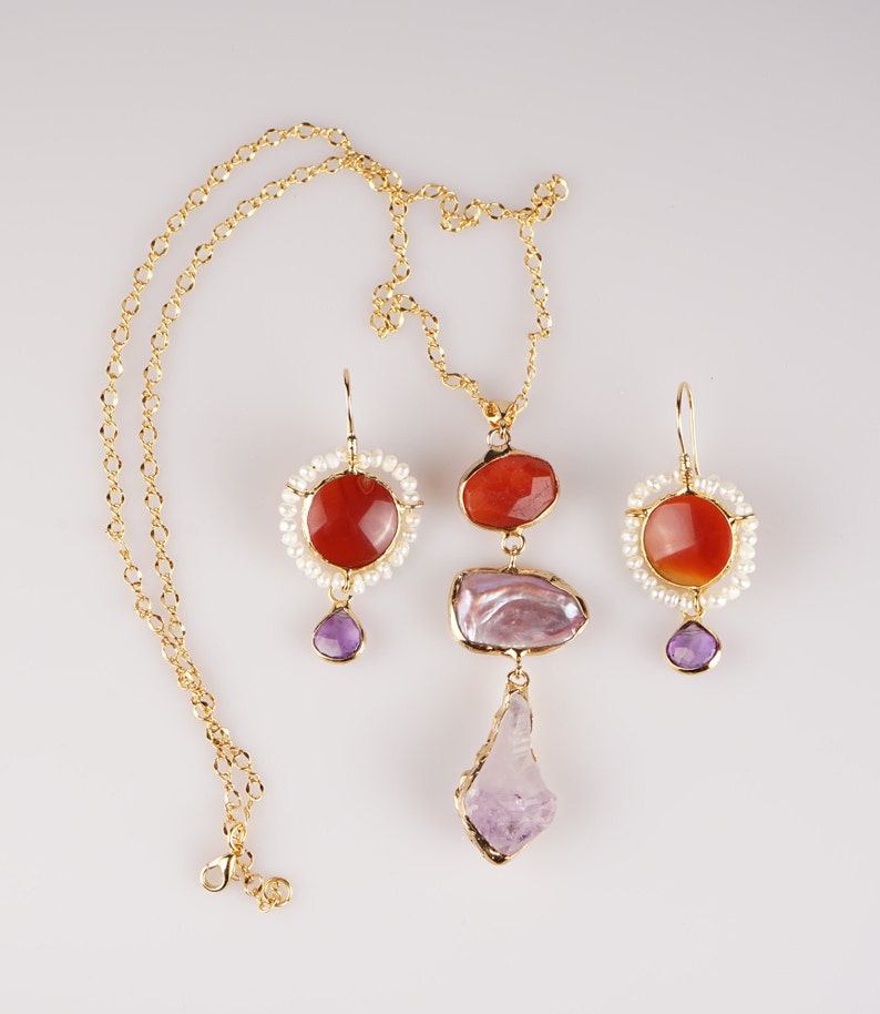 May include: A gold necklace and earrings set with red and purple gemstones. The necklace features a large, irregular-shaped purple stone, a smaller red stone, and a circle of white pearls. The earrings have a similar design with a red stone surrounded by white pearls and a smaller purple stone dangling below.