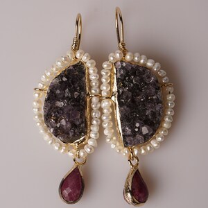 Peut inclure: Une paire de boucles d'oreilles en or avec une pierre violette et une pierre rouge qui pendent. Les boucles d'oreilles sont entourées de perles blanches.
