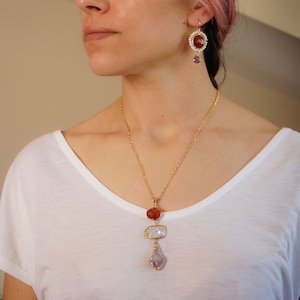 May include: A gold necklace with a pendant featuring three stones: a red carnelian, a white mother-of-pearl, and a purple amethyst. The necklace is paired with matching earrings with a red carnelian stone and a purple amethyst dangle.