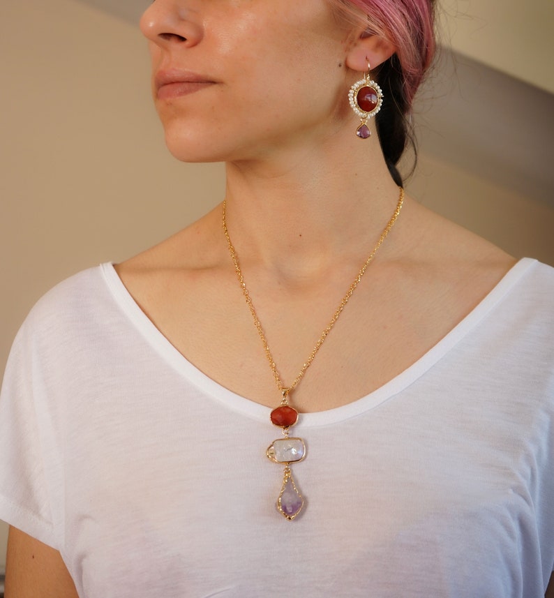 May include: A gold necklace with a pendant featuring three stones: a red carnelian, a white mother-of-pearl, and a purple amethyst. The necklace is paired with matching earrings with a red carnelian stone and a purple amethyst dangle.