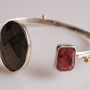 May include: A silver bangle bracelet with two gemstone accents. The larger stone is a dark gray oval shape, and the smaller stone is a rectangular pink stone.