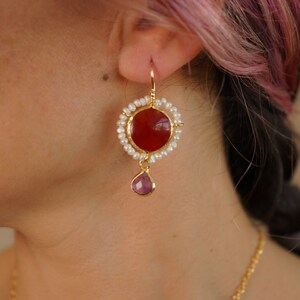 May include: Gold earrings with a red carnelian stone surrounded by white pearls and a dangling purple stone.