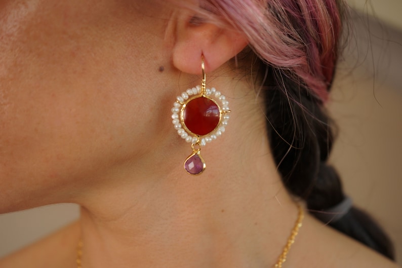 May include: Gold earrings with a red carnelian stone surrounded by white pearls and a dangling purple stone.