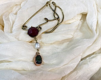 May include: A gold-toned brooch in the shape of a horse's head with a red gemstone and a dangling pearl and green gemstone.