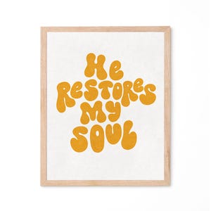May include: A framed print featuring the phrase "He restores my soul" in a retro, bubble-style font. The text is a warm yellow against a white backdrop. The frame is a light wood colour.