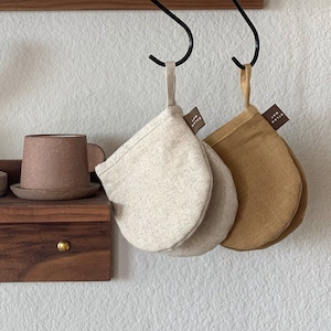 Puede incluir: Exhibición de utensilios de cocina en una estantería de madera. La estantería contiene varias tazas, platillos y cuencos de cerámica en tonos neutros. Debajo, cuelgan dos manoplas para horno en beige y marrón claro. La estantería está montada en una pared blanca.