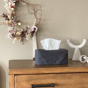 May include: A dark gray tissue box cover with white tissues peeking out, sits on a wooden dresser. A decorative wreath with dried flowers and berries hangs on the wall. A white, curved sculpture is also on the dresser.
