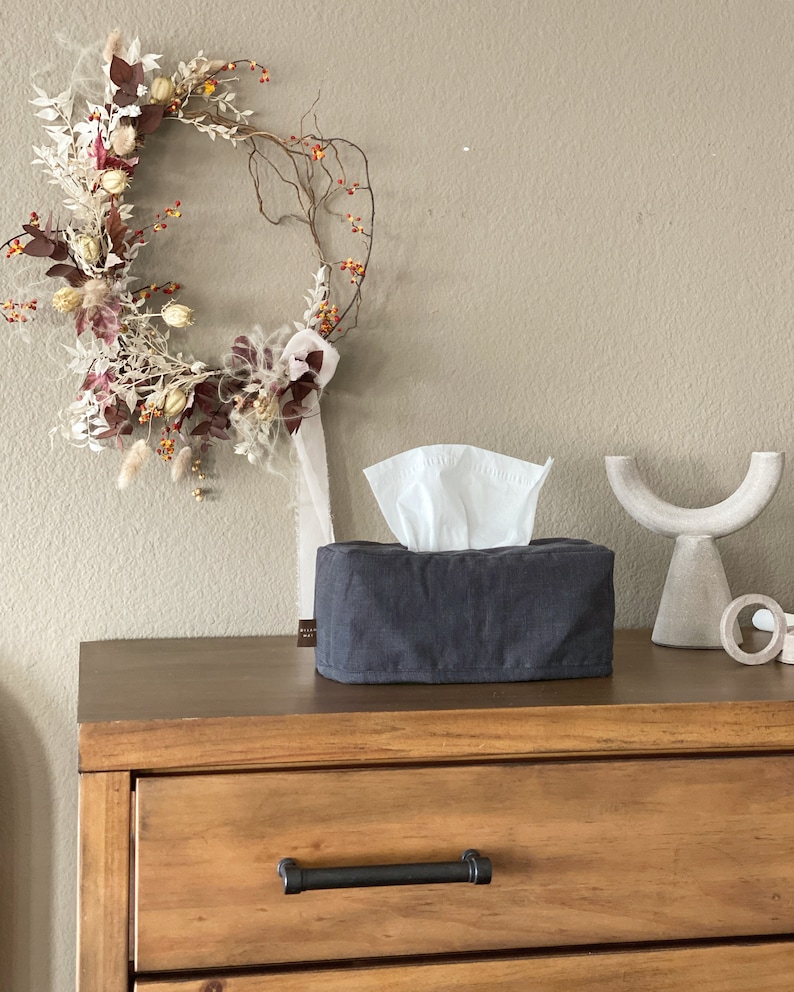 May include: A dark gray tissue box cover with white tissues peeking out, sits on a wooden dresser. A decorative wreath with dried flowers and berries hangs on the wall. A white, curved sculpture is also on the dresser.