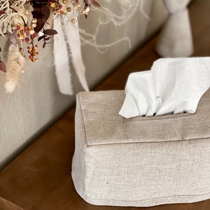 May include: A rectangular tissue box cover made of light beige fabric, with a stack of white tissues protruding from the top. The box sits on a wooden surface, with dried floral arrangements and a decorative candle holder in the background.
