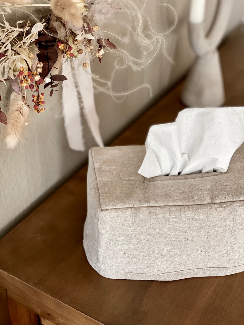 May include: A rectangular tissue box cover made of light beige fabric, with a stack of white tissues protruding from the top. The box sits on a wooden surface, with dried floral arrangements and a decorative candle holder in the background.