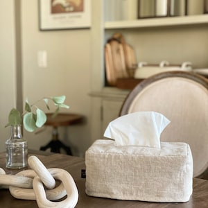 May include: A rectangular tissue box cover made of beige linen fabric with a stack of white tissues emerging from the top. A decorative wooden chain and a small glass vase with greenery are also on the table.