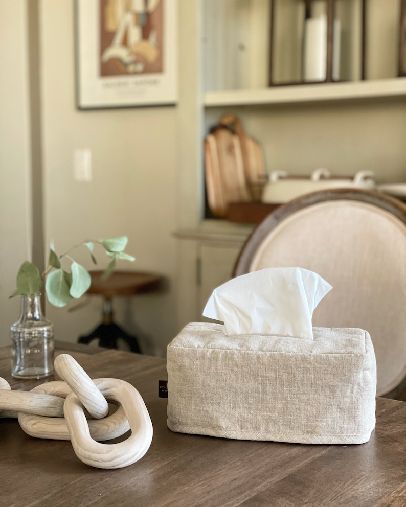 May include: A rectangular tissue box cover made of beige linen fabric with a stack of white tissues emerging from the top. A decorative wooden chain and a small glass vase with greenery are also on the table.
