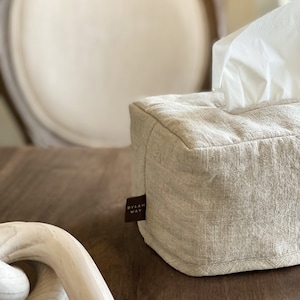 May include: A rectangular tissue box cover made of beige linen fabric. White tissues are visible at the top. A small brown tag with the text "DYLAN WAY" is attached to the side. The box is on a wooden surface.