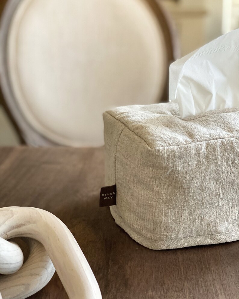 May include: A rectangular tissue box cover made of beige linen fabric. White tissues are visible at the top. A small brown tag with the text "DYLAN WAY" is attached to the side. The box is on a wooden surface.