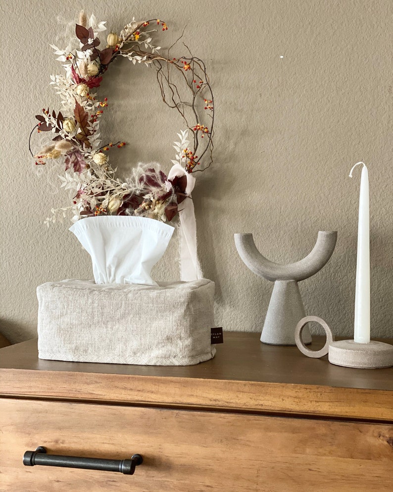 May include: A neutral-toned linen tissue box with a white tissue insert, a decorative wreath with dried flowers, and a modern gray candle holder and a white candle. The items are arranged on a wooden surface.