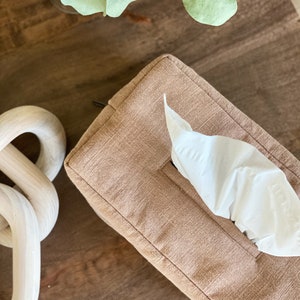 May include: A rectangular tissue box cover in a light brown fabric with a single white tissue protruding from the opening. A decorative wooden chain and sprigs of greenery are also visible.